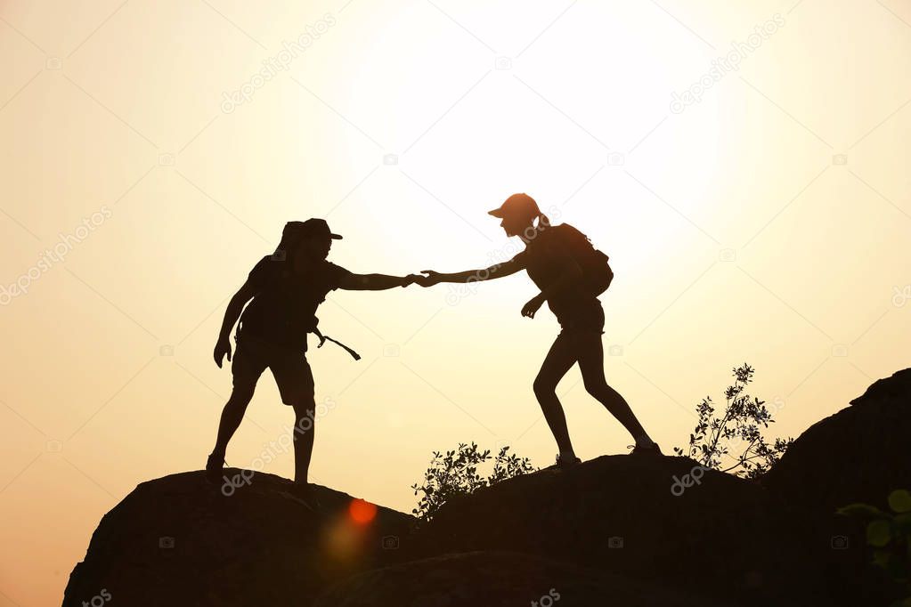 Silhouettes of man and woman helping each other to climb on hill against sunset