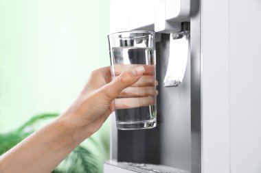 Woman filling glass from water cooler indoors, closeup. Refreshing drink clipart