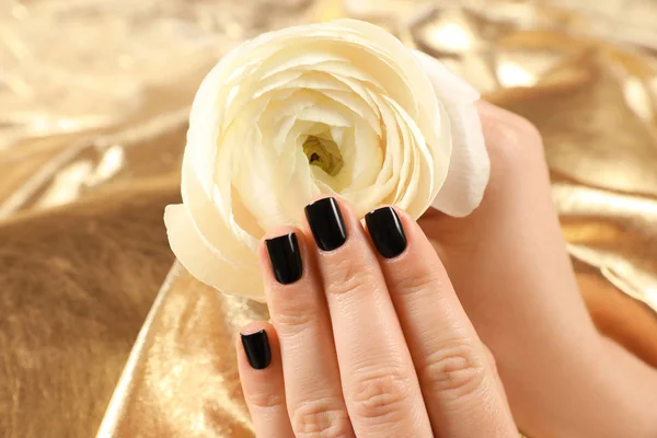 Mujer con manicura negra sosteniendo hermosa flor sobre fondo dorado, primer plano. Tendencias de esmalte de uñas —  Fotos de Stock
