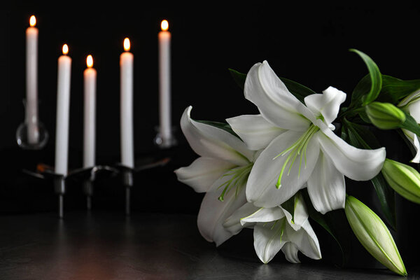 Lírios brancos e velas borradas acesas na mesa na escuridão, fechando com espaço para texto. Símbolo funerário — Fotografia de Stock
