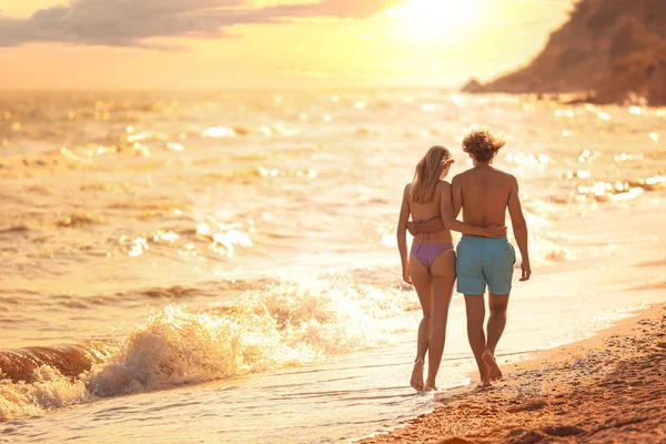 Młoda kobieta w bikini i jej chłopak chodzenie na plaży o zachodzie słońca. Piękna para — Zdjęcie stockowe