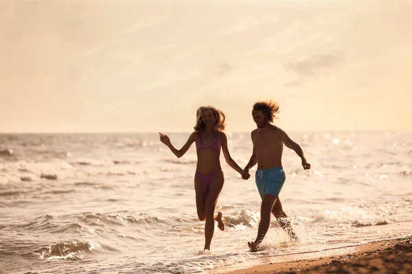 穿着比基尼的年轻女子和她的男朋友在日落时分在沙滩上玩得很开心。可爱的夫妇 — 图库照片
