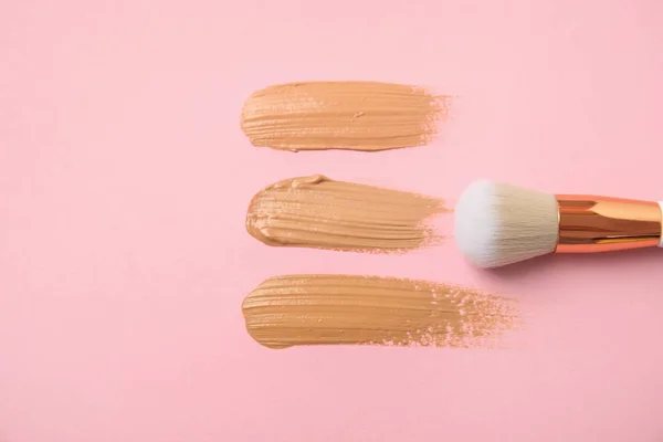 Samples of different foundation shades and makeup brush on pink background, above view