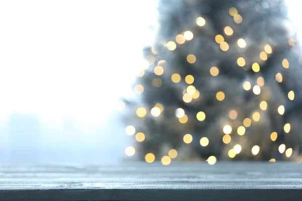 Vista borrosa de hermoso árbol de Navidad con luces amarillas cerca de la ventana interior, se centran en la mesa de madera. Espacio para texto — Foto de Stock