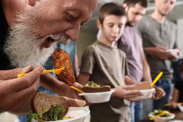 Hombre mayor con otras personas pobres comiendo comida en el centro de caridad — Foto de Stock