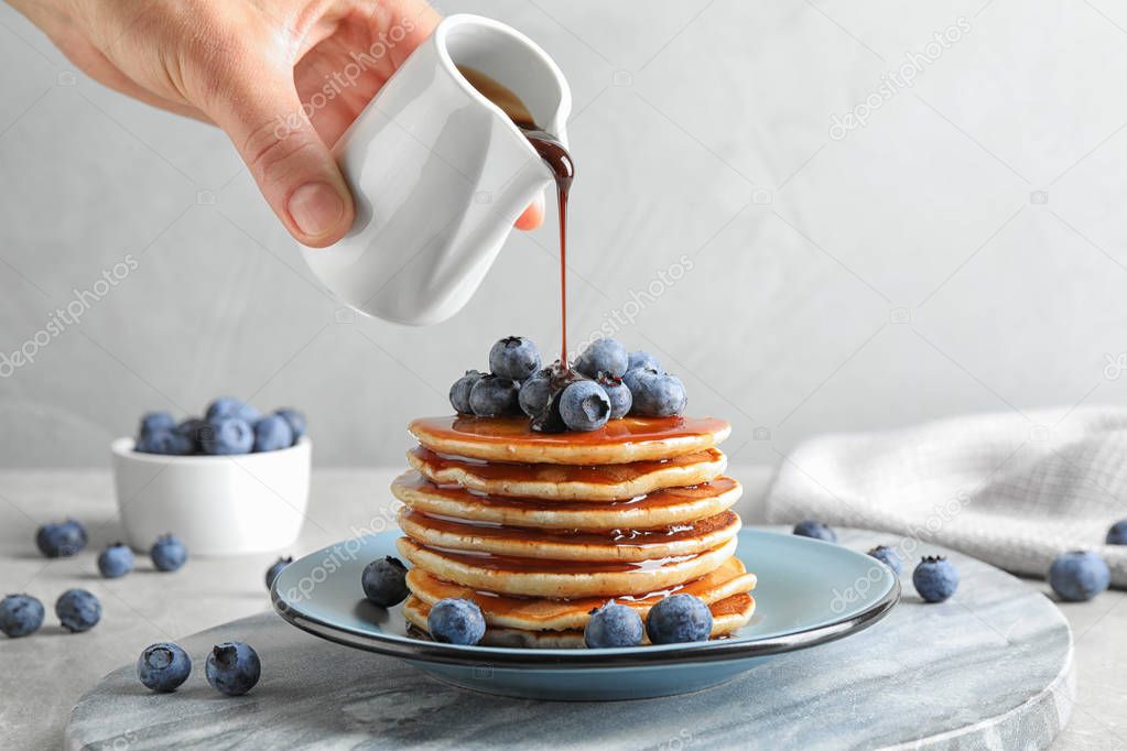 Woman pouring chocolate syrup onto fresh pancakes with blueberries at grey table, closeup