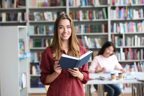 Jovem mulher bonita com livro na biblioteca — Fotografia de Stock