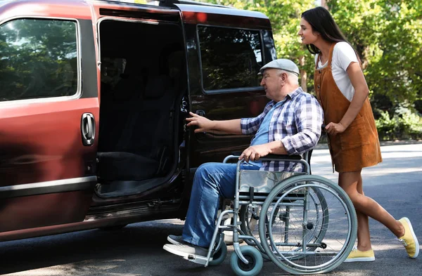 Молодая женщина помогает пожилому мужчине в инвалидной коляске сесть в фургон на улице — стоковое фото