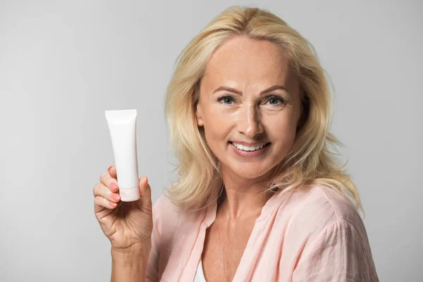 Retrato de hermosa mujer madura con piel perfecta sosteniendo tubo de crema sobre fondo gris —  Fotos de Stock