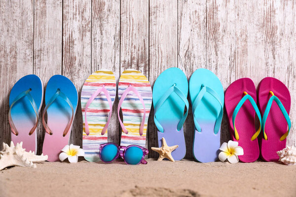 Different bright flip flops and sunglasses on sand near wooden wall, space for text. Summer beach accessories