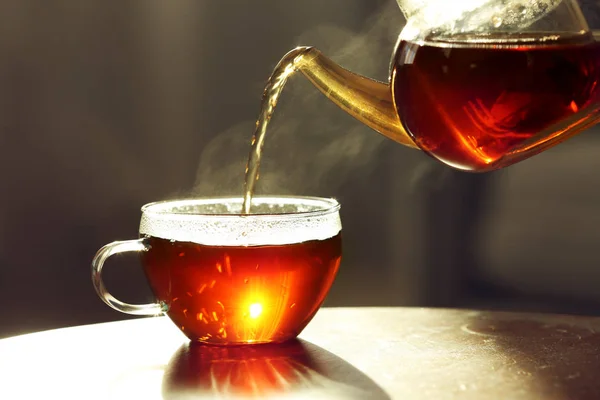 Pouring fresh hot tea from teapot into cup against blurred background, closeup — Stock Photo, Image