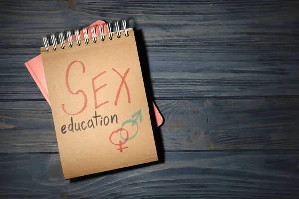 Cuaderno con frase "SEXO EDUCACIÓN" y símbolos de género sobre fondo de madera oscura, vista superior. Espacio para texto — Foto de Stock