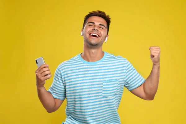 Joven feliz con smartphone escuchando música a través de auriculares inalámbricos sobre fondo amarillo — Foto de Stock