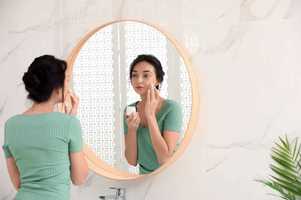 Giovane donna che applica la crema sul viso vicino specchio in bagno — Foto Stock