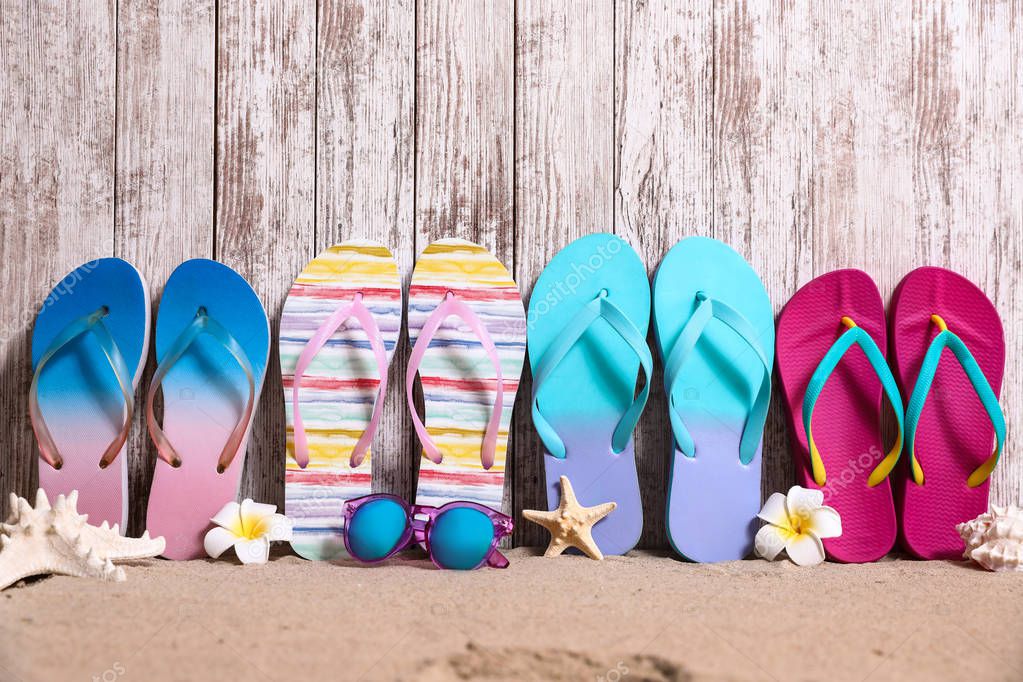 Different bright flip flops and sunglasses on sand near wooden wall, space for text. Summer beach accessories
