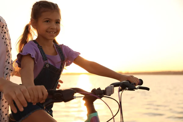 Szczęśliwy matka teaching jej córka do jeździć rower w pobliżu rzeka w zachód słońca — Zdjęcie stockowe