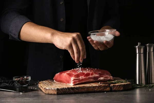 Uomo salatura carne fresca cruda sul tavolo contro sfondo scuro, primo piano — Foto Stock