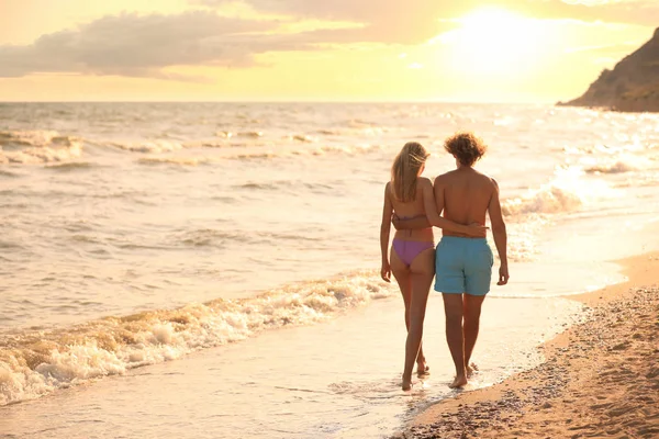 Jeune femme en bikini et son petit ami marchant sur la plage au coucher du soleil. Charmant couple — Photo