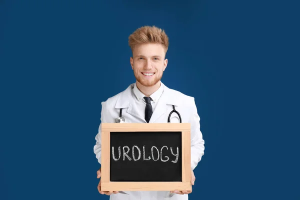 Homme médecin tenant petit tableau noir avec mot UROLOGIE sur fond bleu — Photo