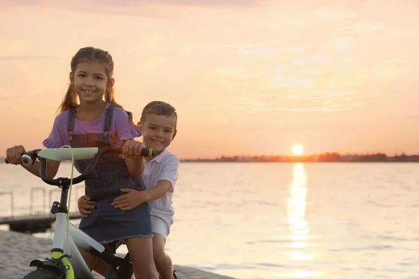 Šťastné malé sourozence jezdiající na kole u řeky při západu slunce — Stock fotografie