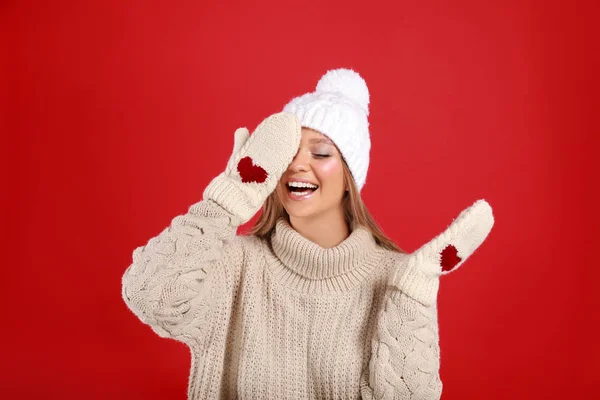 Rolig ung kvinna i varm tröja, tumvantar och mössa på röd bakgrund. Vintersäsongen — Stockfoto