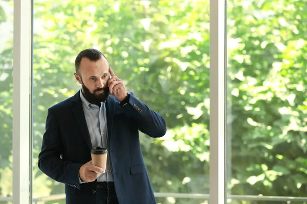 Portret van volwassen man in elegante pak met koffie praten op mobiele telefoon in de buurt van venster. ruimte voor tekst — Stockfoto