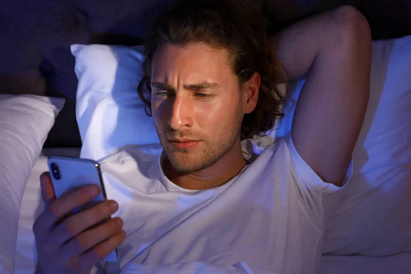 Красивый молодой человек, использующий смартфон, пока лежит на подушке ночью, вид сверху. Время спать — стоковое фото