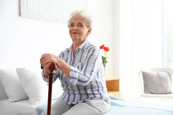Elderly woman with walking cane sitting on bed in nursing home. Medical assistance