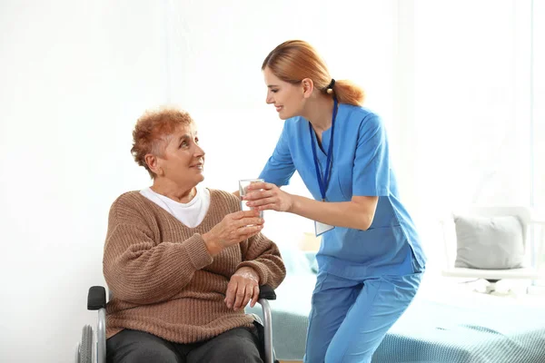 Enfermera dando vaso de agua a anciana en silla de ruedas en interiores. Asistencia médica — Foto de Stock