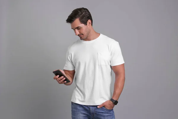 Pohledný mladý muž se smartphone na šedém pozadí — Stock fotografie