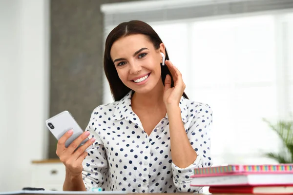 Mujer joven feliz con teléfono inteligente escuchando música a través de auriculares inalámbricos en la mesa en la oficina — Foto de Stock