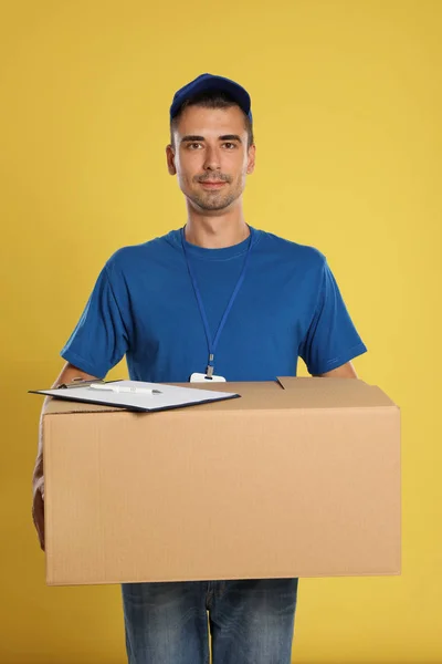 Happy young courier with cardboard box and clipboard on yellow background — Stok Foto