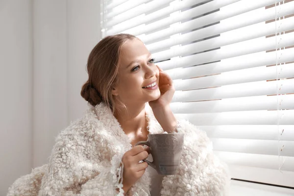 Krásná mladá žena s šálkem teplého nápoje blízko okna doma. Zimní atmosféra — Stock fotografie