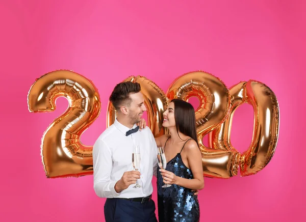 Feliz pareja joven con copas de champán cerca de globos dorados 2020 sobre fondo rosa. Celebración de Año Nuevo — Foto de Stock