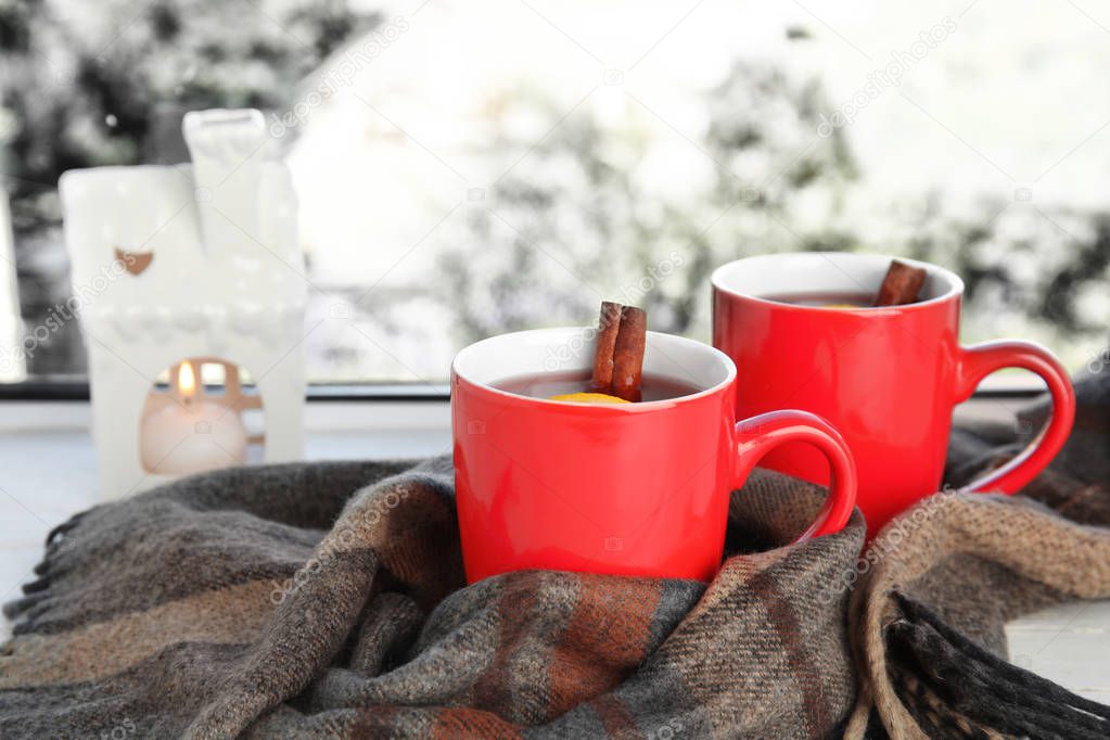 Cups of hot winter drink with scarf on window sill indoors