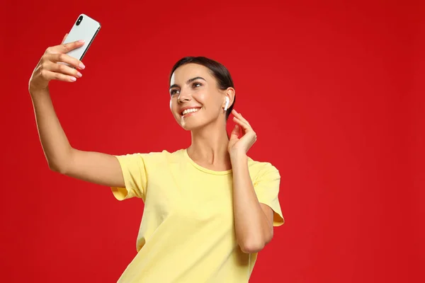Jovem feliz com fones de ouvido sem fio tirando selfie no fundo vermelho — Fotografia de Stock