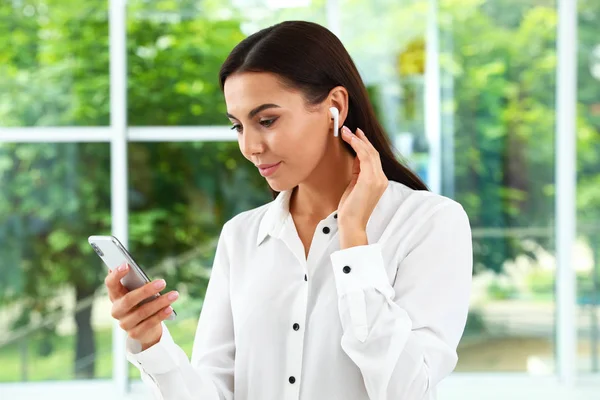 Mujer joven con smartphone escuchando música a través de auriculares inalámbricos en interiores — Foto de Stock