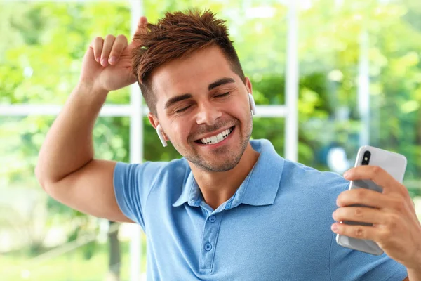 Joven feliz con smartphone escuchando música a través de auriculares inalámbricos en el interior — Foto de Stock