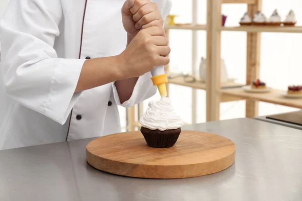 Young female pastry chef decorating cupcake with cream at table in kitchen, closeup