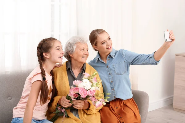 Irmãs felizes tomando selfie com sua avó segurando flores em casa — Fotografia de Stock
