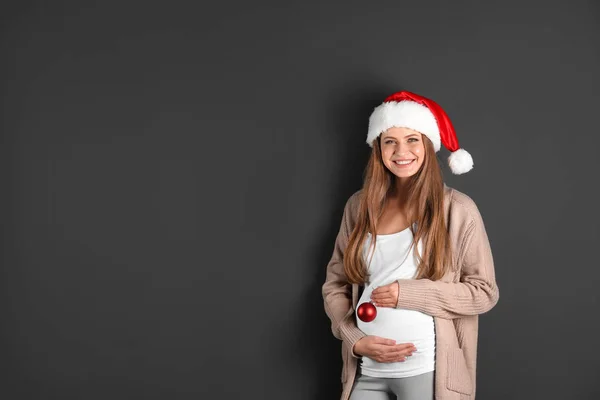灰色の背景にクリスマスの装飾とサンタ帽子の若い妊娠中の女性、テキストのためのスペース — ストック写真