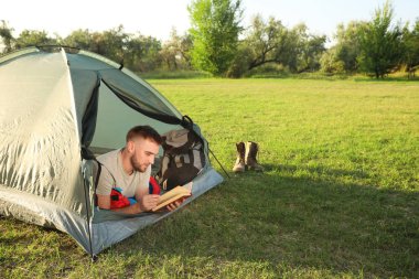 Uyku tulumunda kitap taşıyan genç bir adam kamp çadırında yatıyor.
