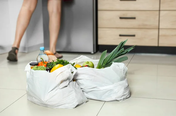 Πλαστικές σακούλες με λαχανικά στο πάτωμα και νέα γυναίκα στην κουζίνα — Φωτογραφία Αρχείου