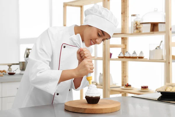 Νεαρή γυναίκα σεφ ζαχαροπλαστικής διακόσμηση cupcake με κρέμα στο τραπέζι στην κουζίνα — Φωτογραφία Αρχείου