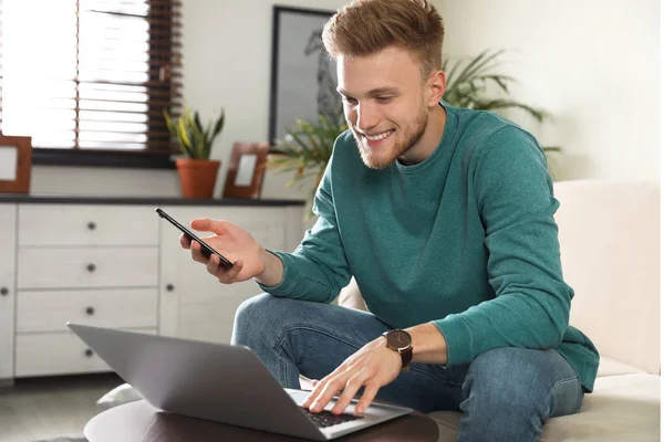 Junger Mann mit Smartphone und Laptop im Wohnzimmer — Stockfoto