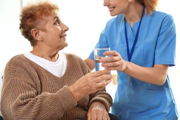 Νοσοκόμα δίνοντας ένα ποτήρι νερό σε ηλικιωμένη γυναίκα σε εσωτερικούς χώρους. Ιατρική βοήθεια — Φωτογραφία Αρχείου