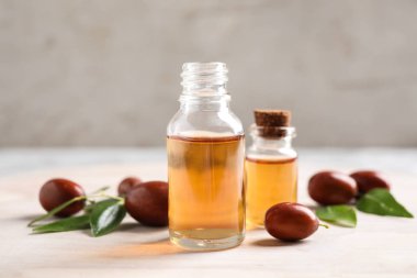 Glass bottles with jojoba oil and seeds on light table against grey background clipart