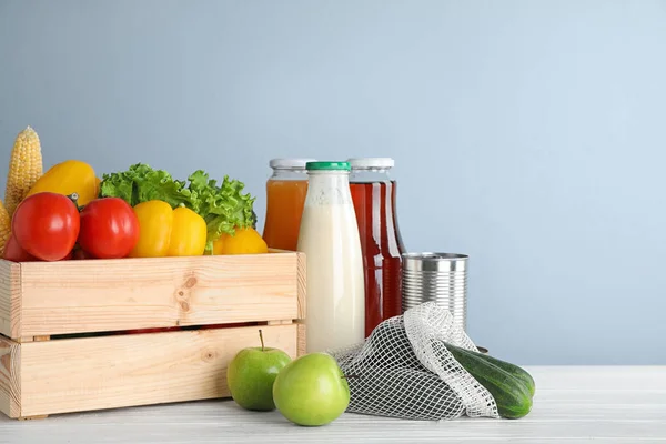 Cassa con verdure fresche e altri prodotti su tavolo di legno bianco su sfondo azzurro — Foto Stock