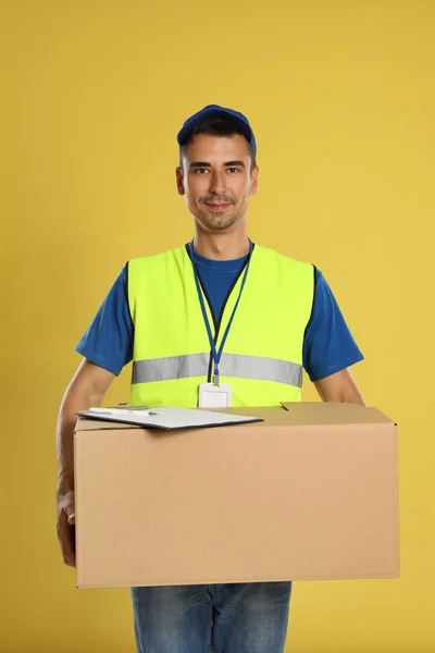Happy young courier with cardboard box and clipboard on yellow background — Stok Foto