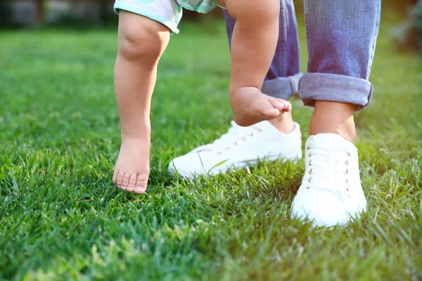 Χαριτωμένο μικρό μωρό που μαθαίνει να περπατήσει με τη νταντά του σε πράσινο γρασίδι σε εξωτερικούς χώρους, κοντινό πλάνο — Φωτογραφία Αρχείου